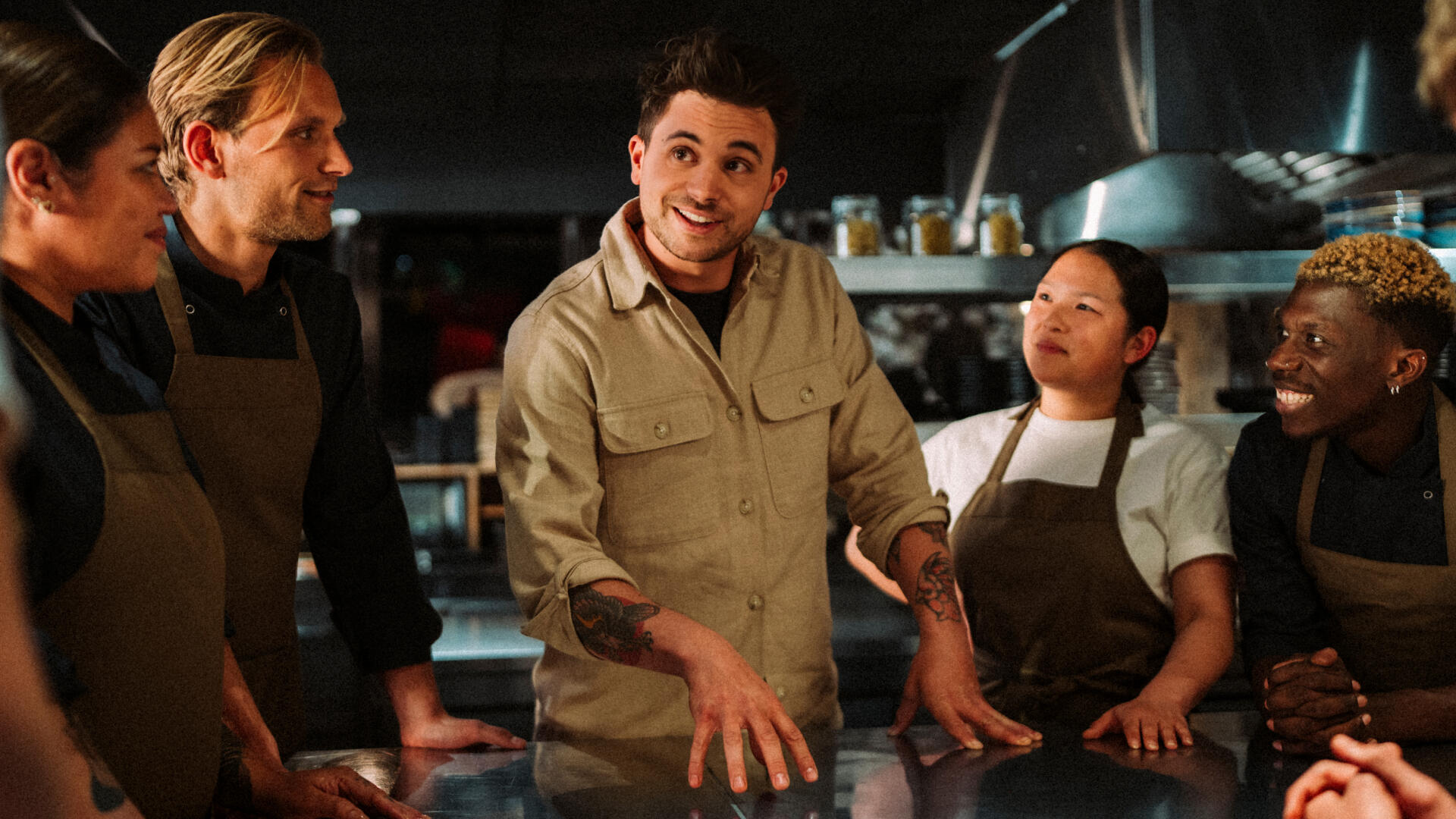 Een team van keukenmedewerkers staat in een halve kring rondom een collega die enthousiast iets uitlegt aan het team.