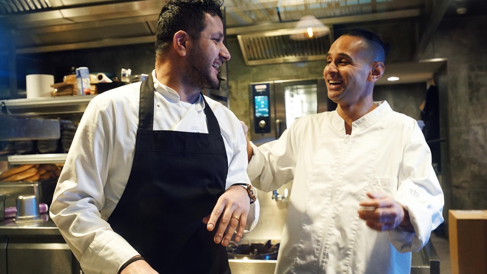 Twee chefs in witte koksbuizen lachen en geven elkaar een vriendschappelijke schouderklop in een drukke restaurantkeuken.