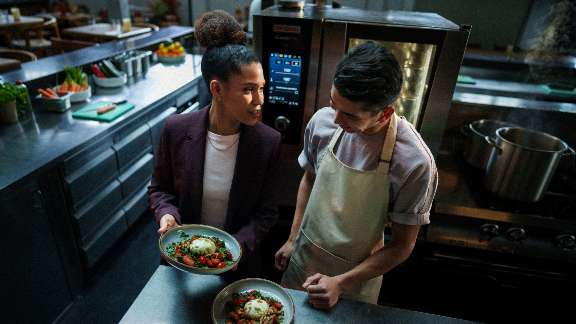 Een vrouwelijke leidinggevende presenteert een mooi opgemaakt bord met burrata en tomaten aan een jonge kok in een industriële keuken. Op de achtergrond is een professionele Rational-oven zichtbaar en de sfeer oogt als een coaching-moment.