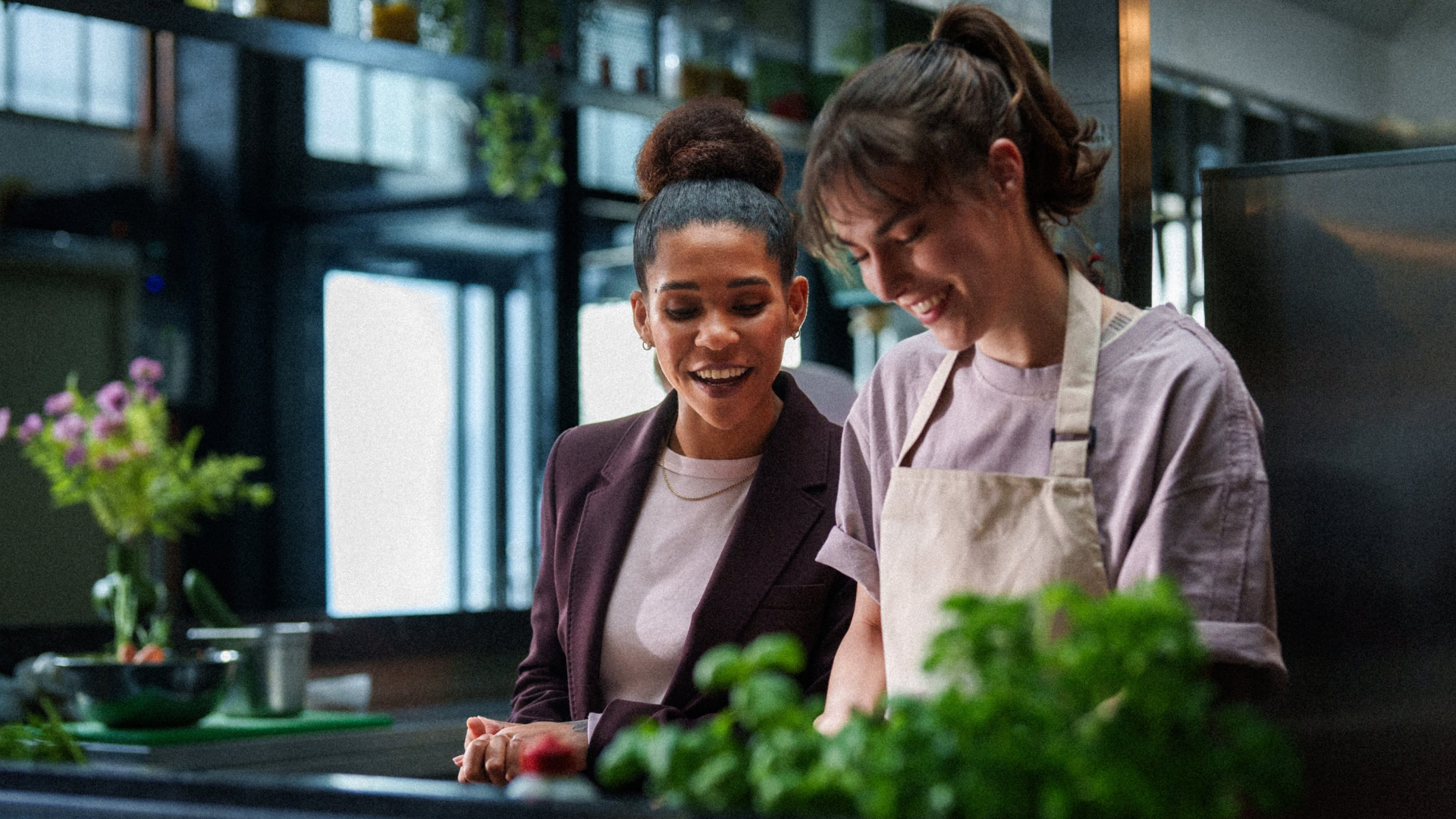 Een glimlachende manager in een bordeauxrood jasje staat naast een medewerker in een beige schort in een professionele keuken. Ze kijken samen naar verse kruiden op het aanrecht in een lichte, moderne horeca-omgeving.