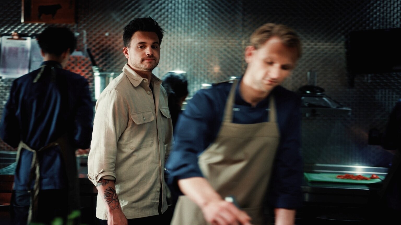 Een man in een beige overhemd observeert het keukenteam tijdens het voorbereiden van gerechten in een professionele keukenomgeving.