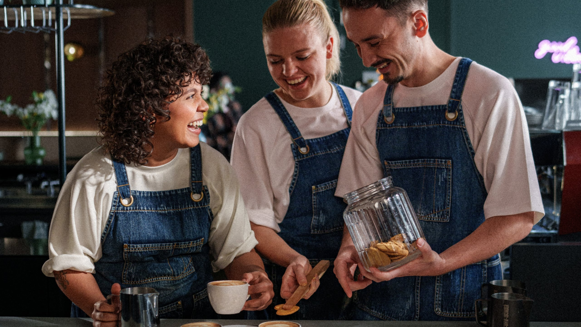 Drie collega's in spijkerstof schorten die samen lachen bij een koffiemachine terwijl een van hen koekjes uit een grote glazen pot pakt.