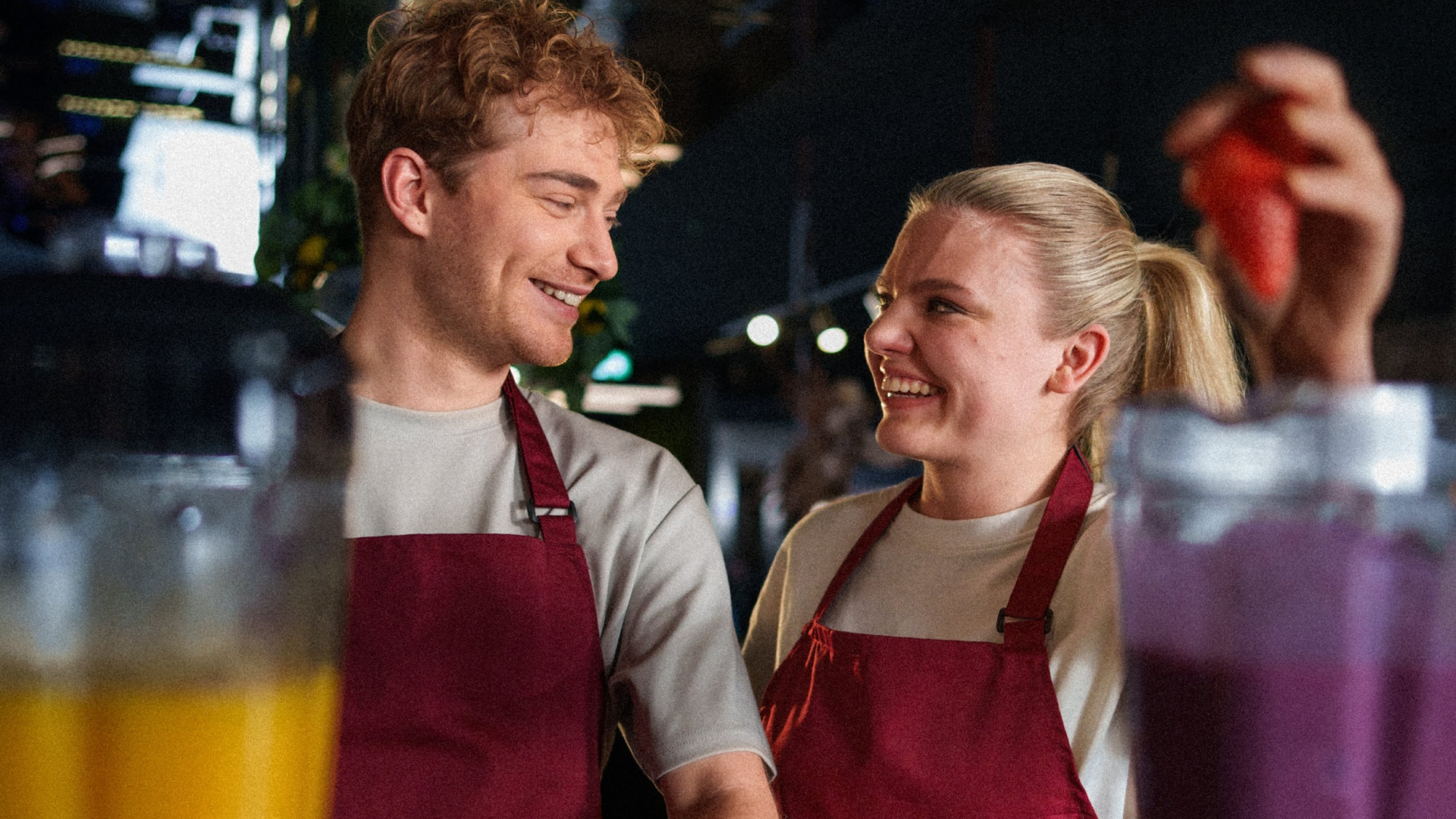 Twee jonge collega's in bedrijfskleding die vrolijk met elkaar praten en lachen terwijl ze samenwerken achter de bar bij een blender.
