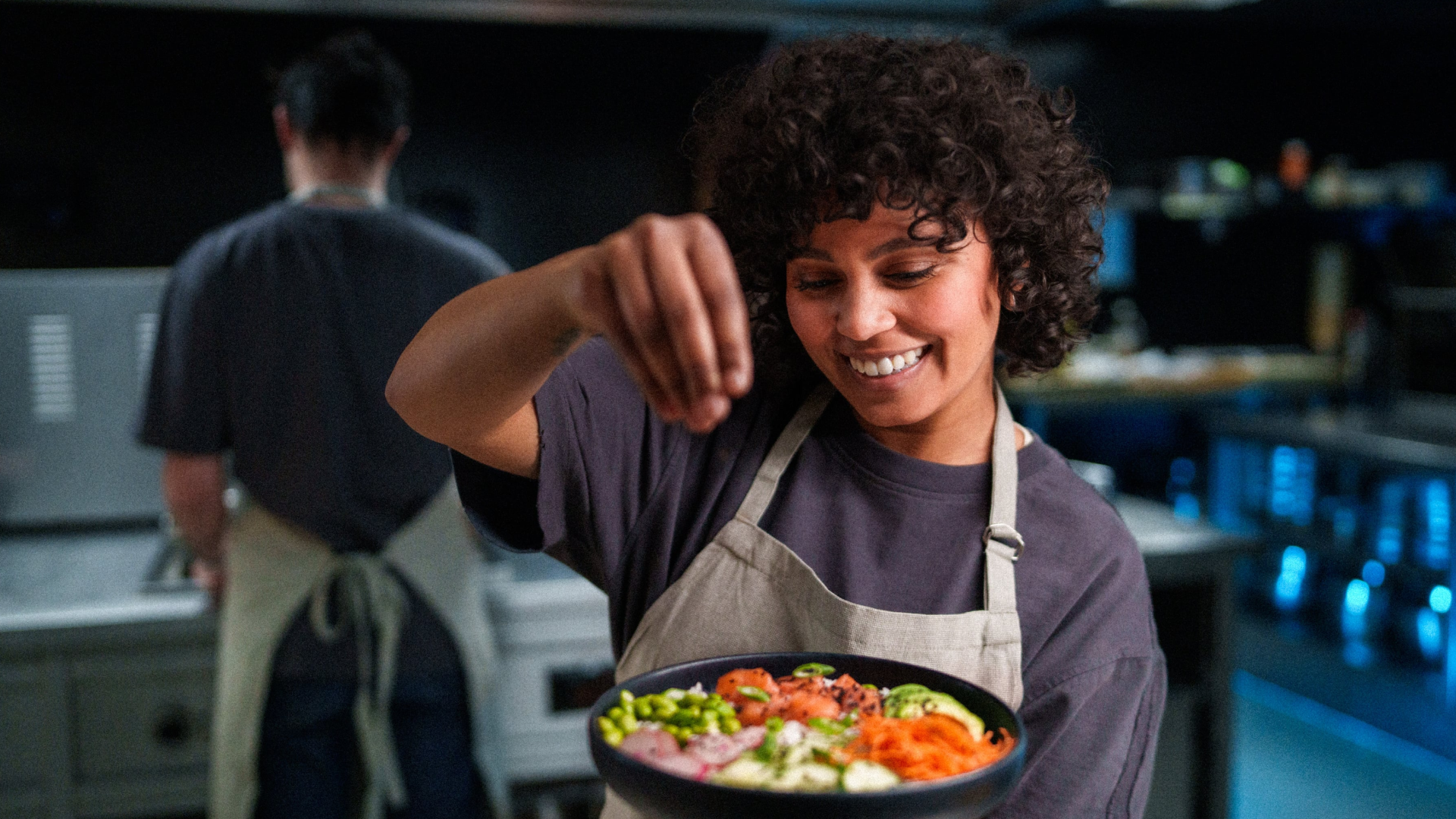 Een geconcentreerde medewerker die met precisie de laatste garnering toevoegt aan een verse, kleurrijke maaltijdsalade in een open keuken.