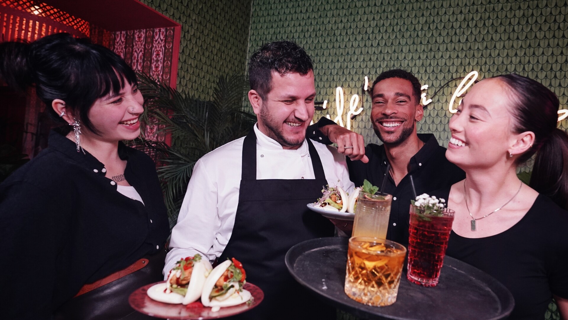 Vier horecamedewerkers lachen en poseren samen in een sfeervol verlichte ruimte, met kleurrijke cocktails en bao buns op hun dienbladen.