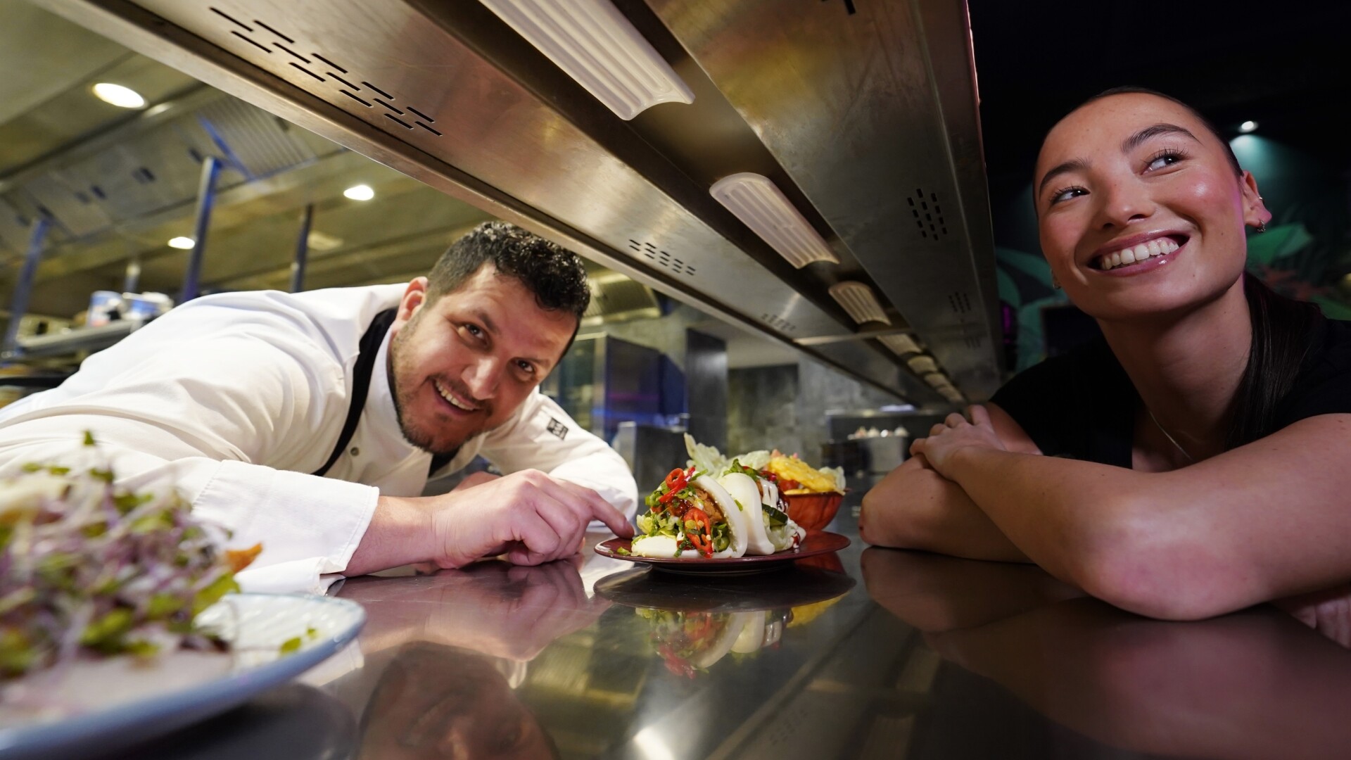 Een chef-kok en een serveerster lachen naar elkaar aan de passe in de keuken, terwijl een kleurrijk gerecht met bao buns net is uitgeserveerd.