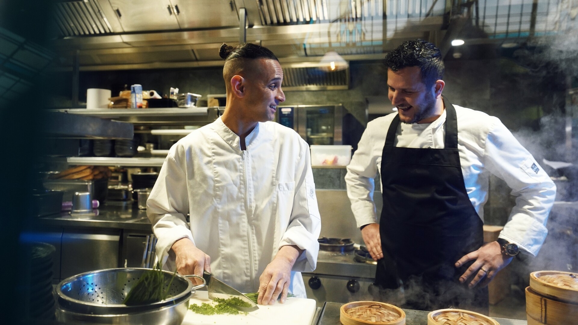 Twee koks in witte jassen staan samen in een professionele keuken, waarbij één kok verse kruiden snijdt en ze elkaar met een glimlach aankijken.